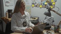 A focused young woman detective with blonde hair works at her office, analyzing evidence on her laptop.