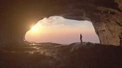 Hiker inside cave looking at sunrise above clouds, adventurous man watching sun rising and shining through clouds. 3D animation.