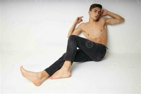 Cute young guy with a naked torso sits on the floor barefoot isolated ...