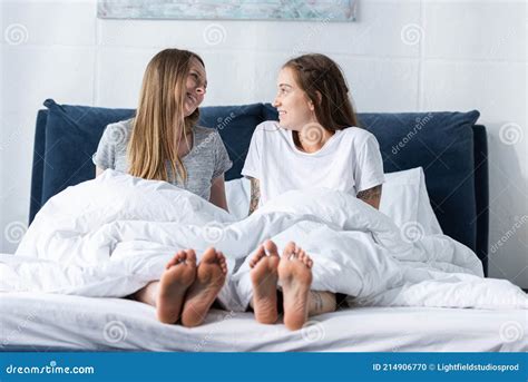 Two Smiling Barefoot Lesbians Sitting on Bed and Looking at Each Other ...