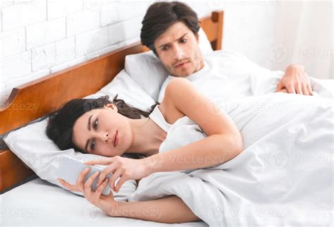 A woman is lying in bed, looking at her phone while a man, who is also ...