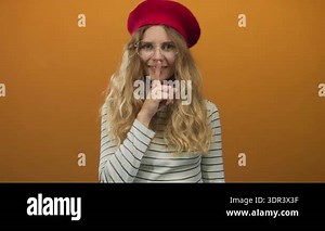 Woman presses finger shushing in a studio featuring blonde model wearing beret and glasses highlighting lips and smile against orange background Stock Video Footage - Alamy
