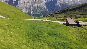 Aerial reveal shot of a hiker walking by a tiny wooden cabin on a hiking trail with view to Snow-capped swiss alp mountains Schreckhorn and Finsteraarhorn in Summer | Premium Stock Video Footage