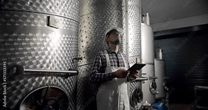 Male wine maker checking quality of beverage distillation process indoors and write down the dates in tablet gadget. Alcohol production concept. Wine production plant. Steel wine distilling tanks.