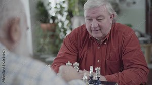 Two mature men playing chess sitting at home. Caucasian old men neighbors friends playing chess joyfully indoors. Intelligence challenge concept