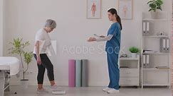Female nurse or caregiver measuring weight on scales of elderly gray-haired woman in clinic and writing down results. Senior patient having consultation during exam in medical office. 4k video.