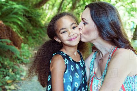 Mother kissing daughter on cheeks against trees - Stock Photo - Dissolve